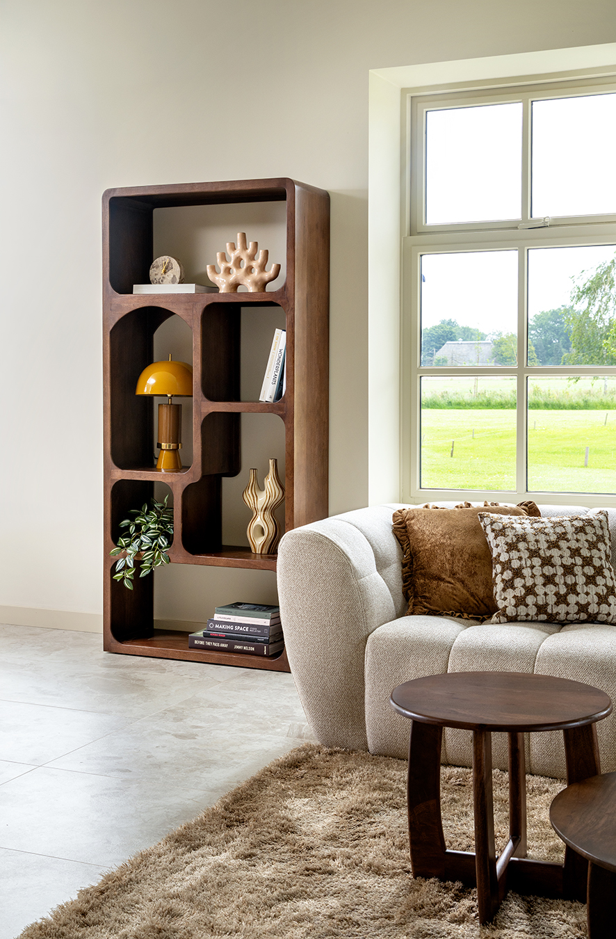 Moderne woonkamer met een organisch gevormde houten kast, een comfortabele crèmekleurige bank, bruine kussens, ronde houten tafels en een raam met uitzicht op groene velden.