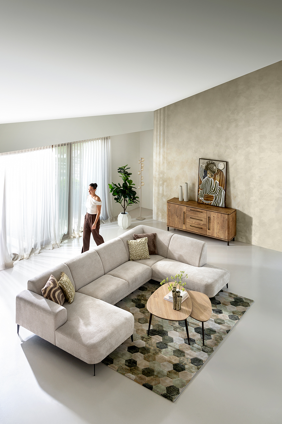 Lichte, moderne woonkamer met een beige hoekbank, een vloerkleed met patroon, houten tafels en een dressoir, grote ramen en een voorbijlopende vrouw.