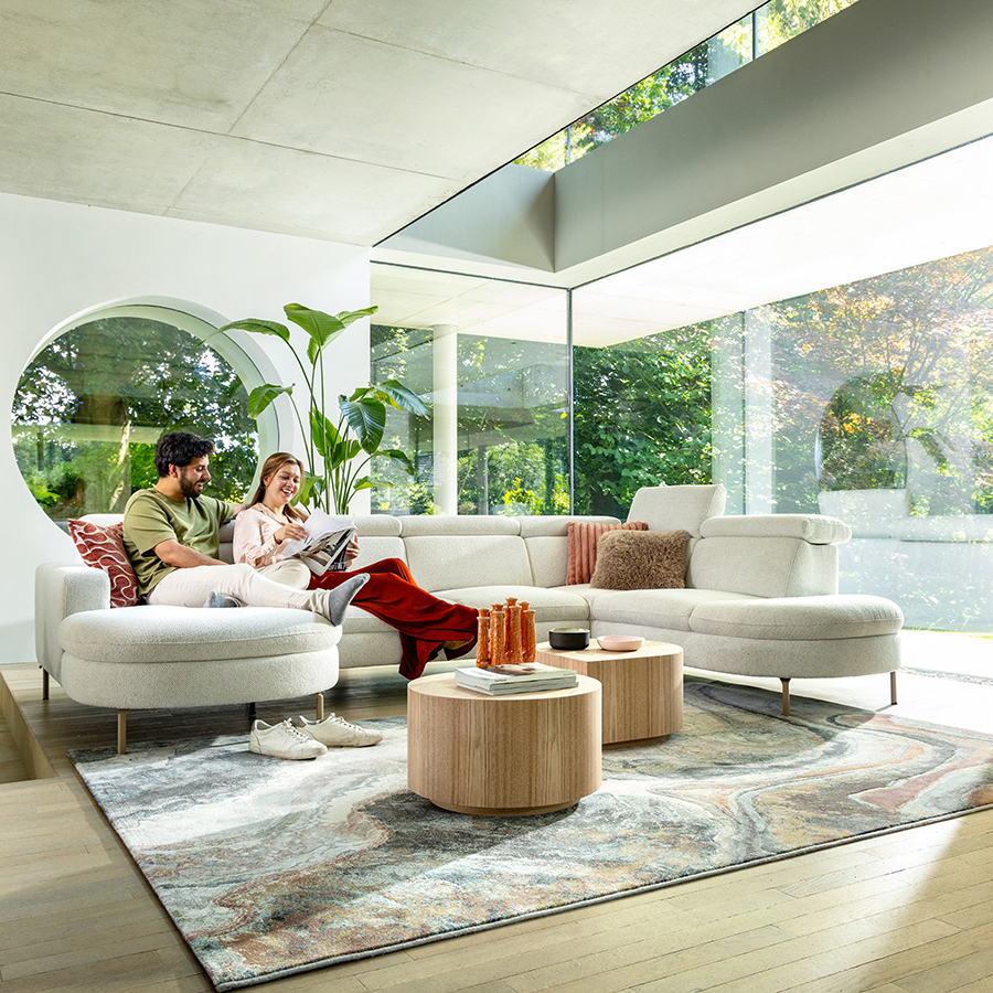 Een stel dat ontspant op een moderne witte hoekbank in een lichte woonkamer met grote ramen, planten en ronde houten salontafels.