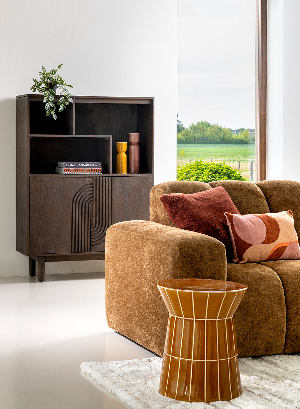 Modern living room with brown sofa, colorful pillows, wooden cabinet, ceramic side table, and large window overlooking green fields