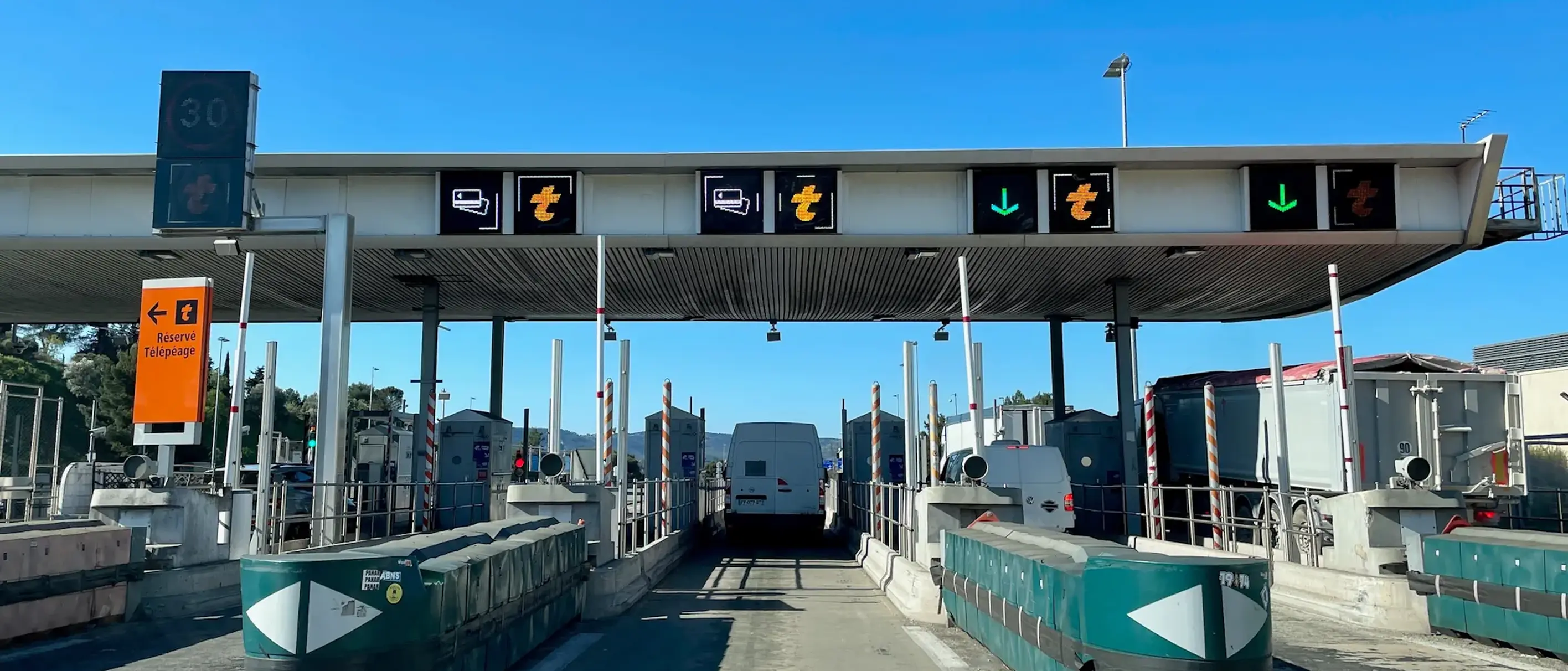 Eine Mautstelle mit mehreren Fahrspuren, Fahrzeuge fahren hindurch. Schilder oben zeigen offene und geschlossene Fahrspuren an. Klarer blauer Himmel im Hintergrund.