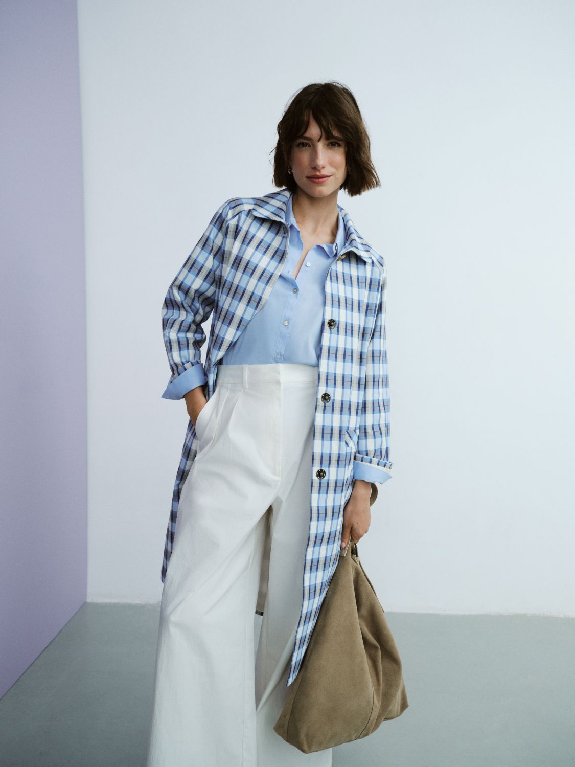 Woman in a blue plaid coat over a light blue shirt and white pants, holding a brown bag, stands against a minimalist backdrop.
