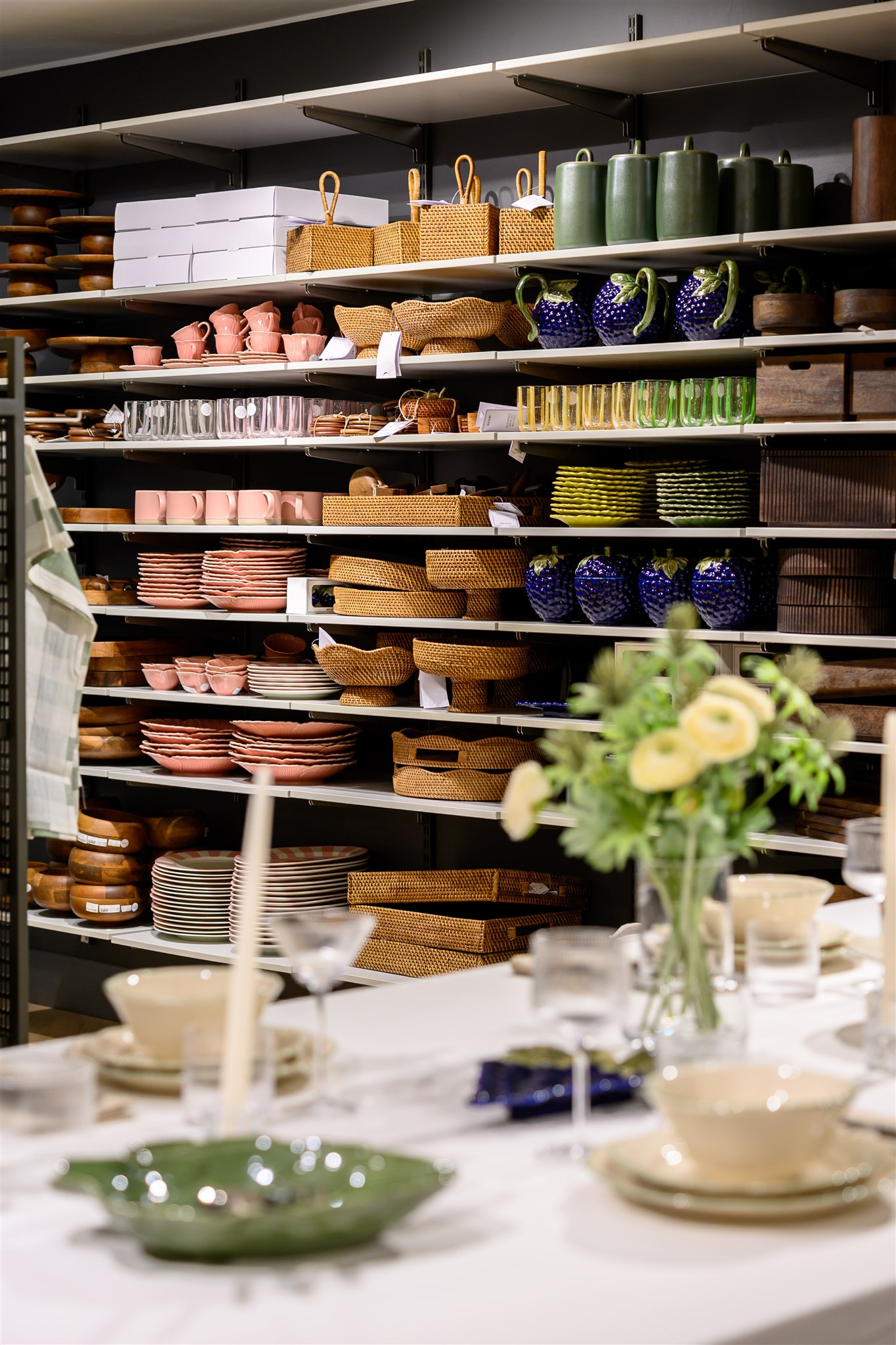 Shelves with neatly arranged ceramics, baskets, and glassware in a store, with a table set in the foreground featuring flowers and dishes.