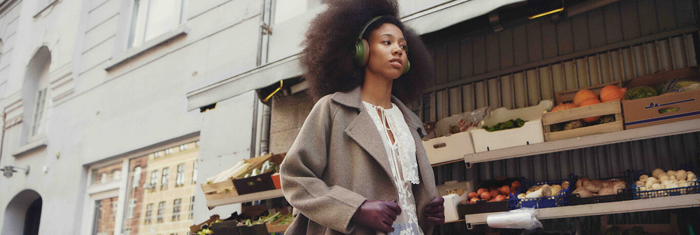 A person wearing headphones and a coat walks past a street market stall with various fruits and vegetables displayed.