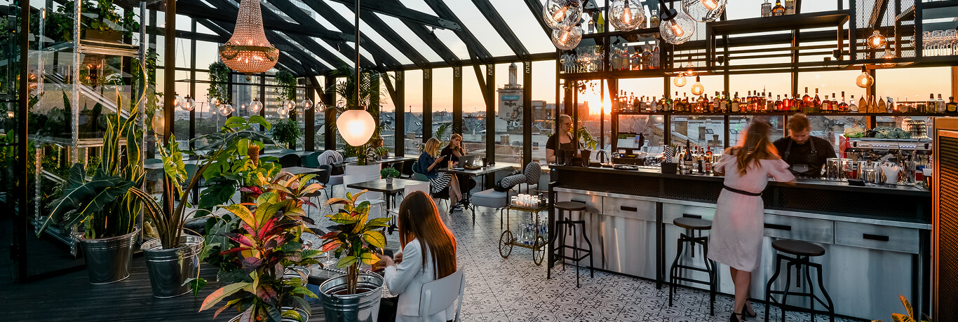 A stylish rooftop bar at sunset with glass walls, lush plants, patrons seated at tables, and a bartender serving drinks.
