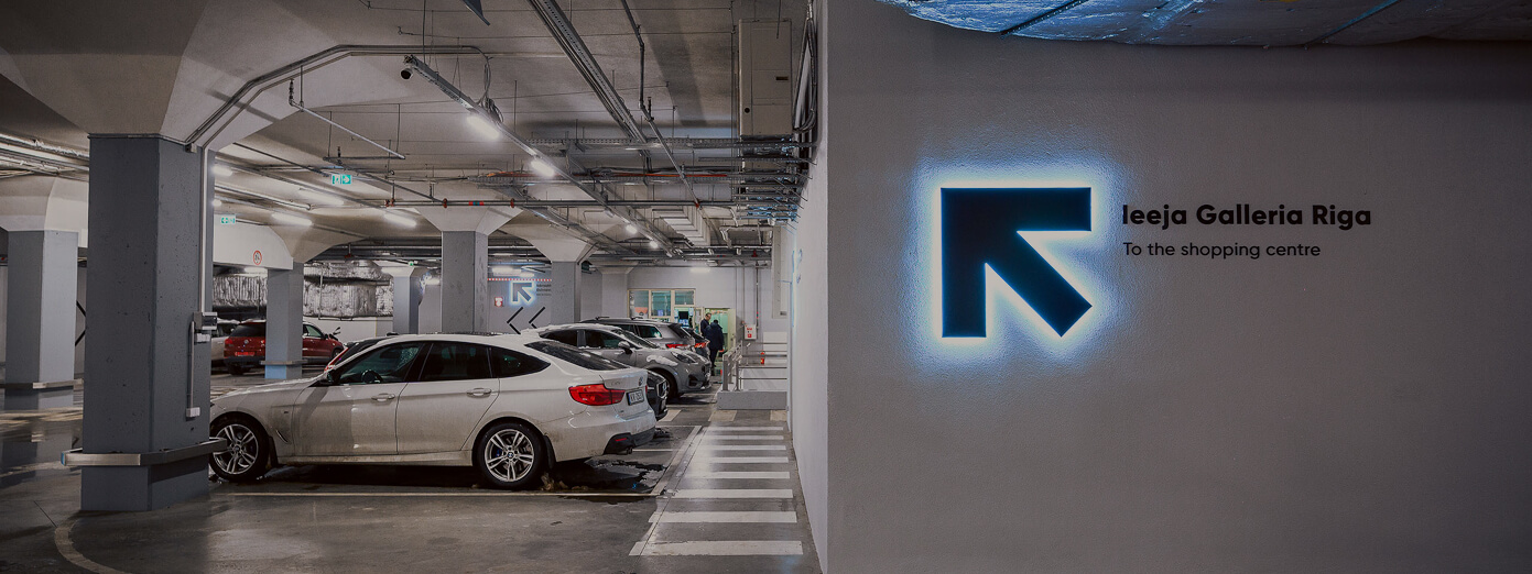 Underground parking garage with several parked cars and a sign pointing towards "Galleria Riga" shopping center.