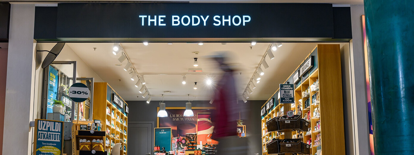 Storefront of The Body Shop with a blurred person walking by. Shelves inside display various products, and a 30% sale sign is visible.