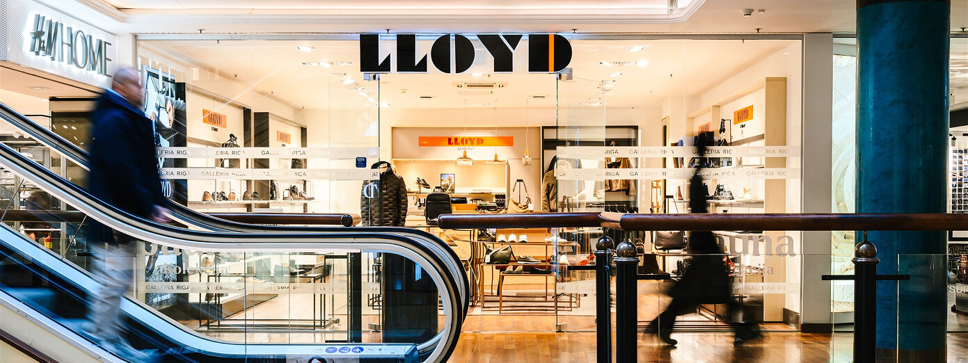 People on an escalator in front of a brightly lit LLOYD shoe store in a shopping mall, displaying various footwear inside.