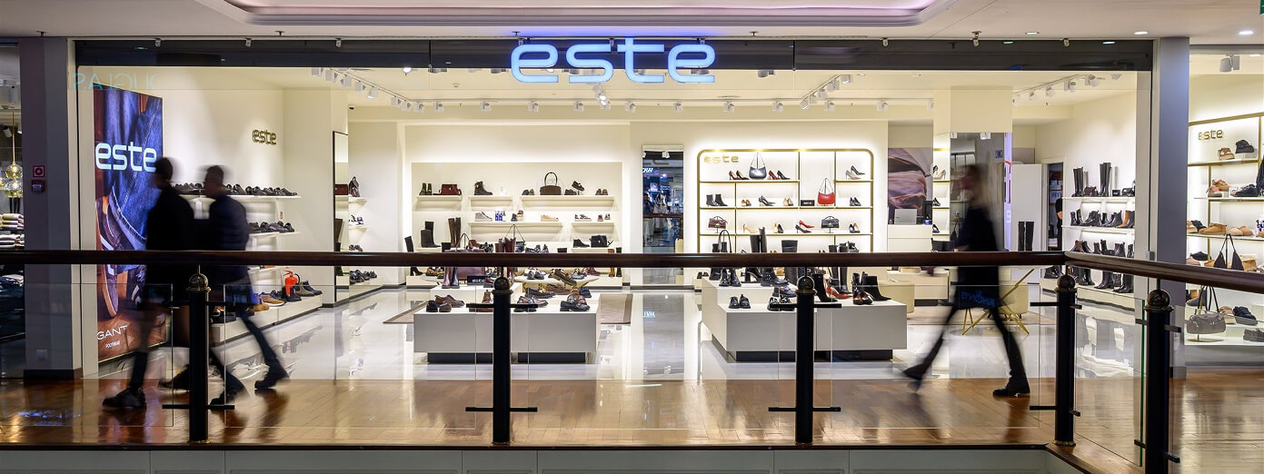 Storefront of a modern shoe and accessory shop named "esté" with illuminated signage, displaying various products inside. People walk past.