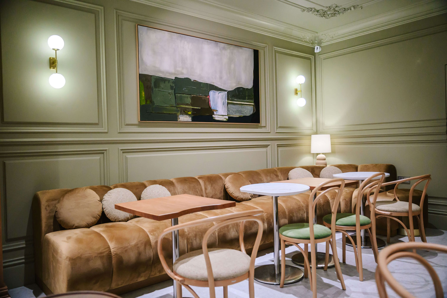 Cozy cafe interior with a brown sofa, round tables, wooden chairs, modern art, and soft lighting on green paneled walls.