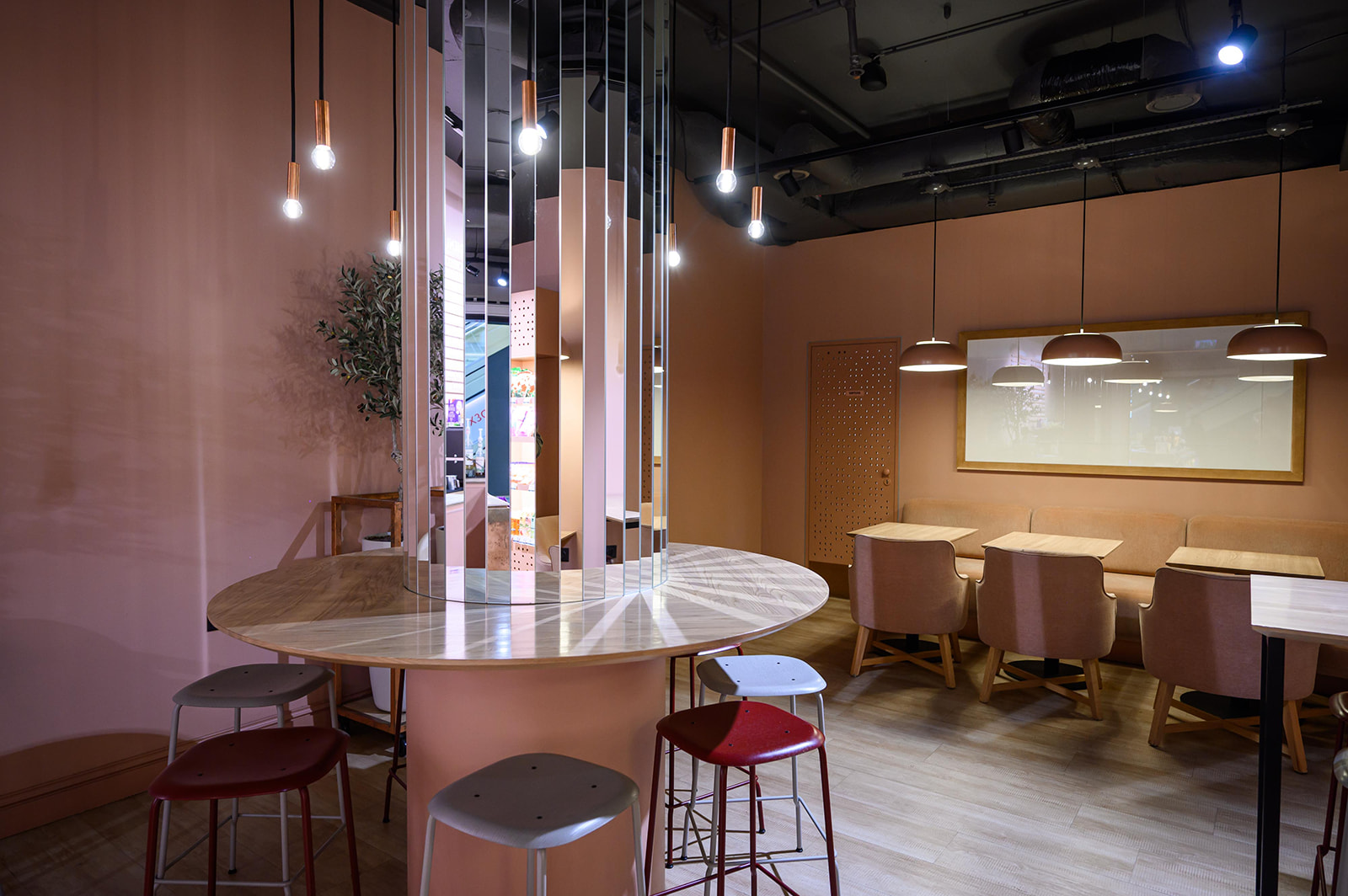 Modern cafe interior with a round table, hanging lights, red stools, and a row of tables with chairs against a peach-colored wall.