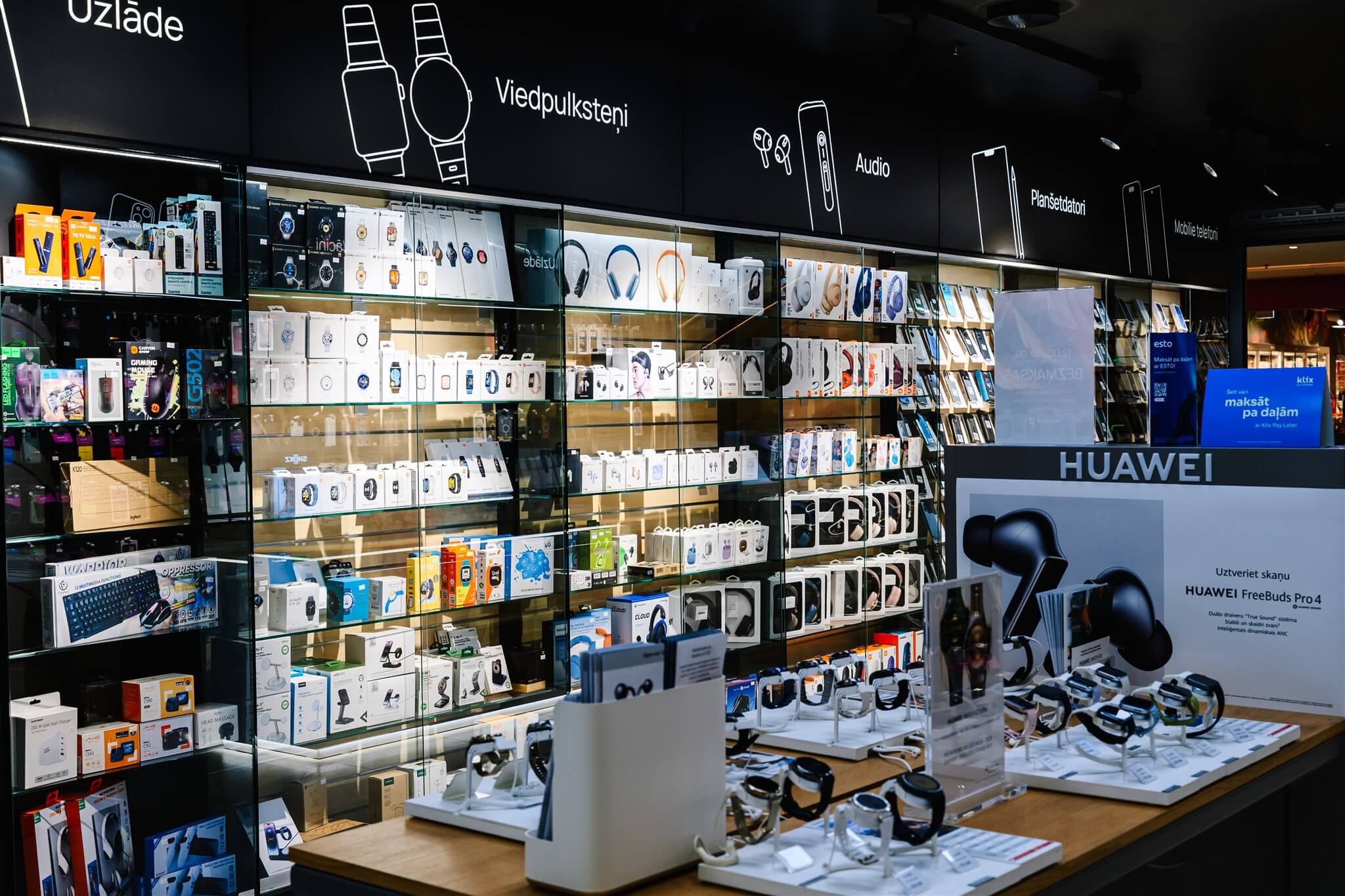 Electronics store display with headphones, smartwatches, and accessories on shelves and tables, under bright lighting.