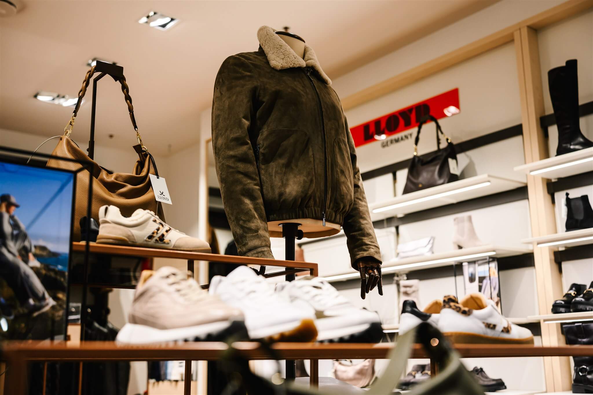 Store display featuring a brown shearling jacket, handbags, and white sneakers, with shelves of shoes and boots in the background.