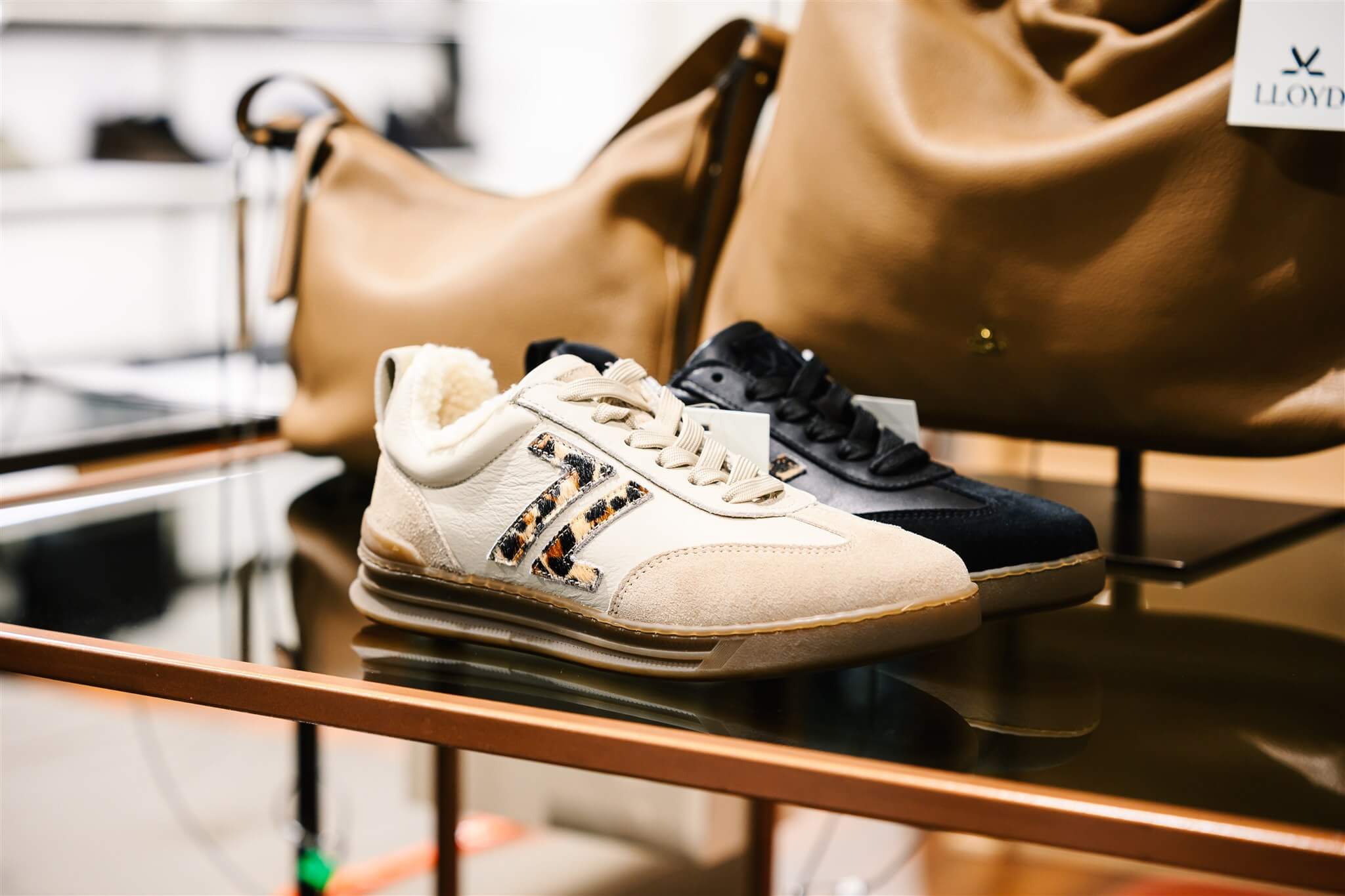 Two pairs of sneakers, one beige with leopard accents and one black, displayed on a glass shelf with a tan handbag in the background.