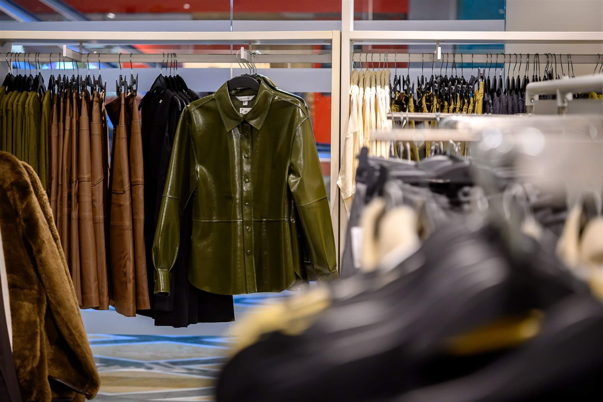 A green leather shirt is prominently displayed on a rack in a clothing store, surrounded by various garments in neutral tones.