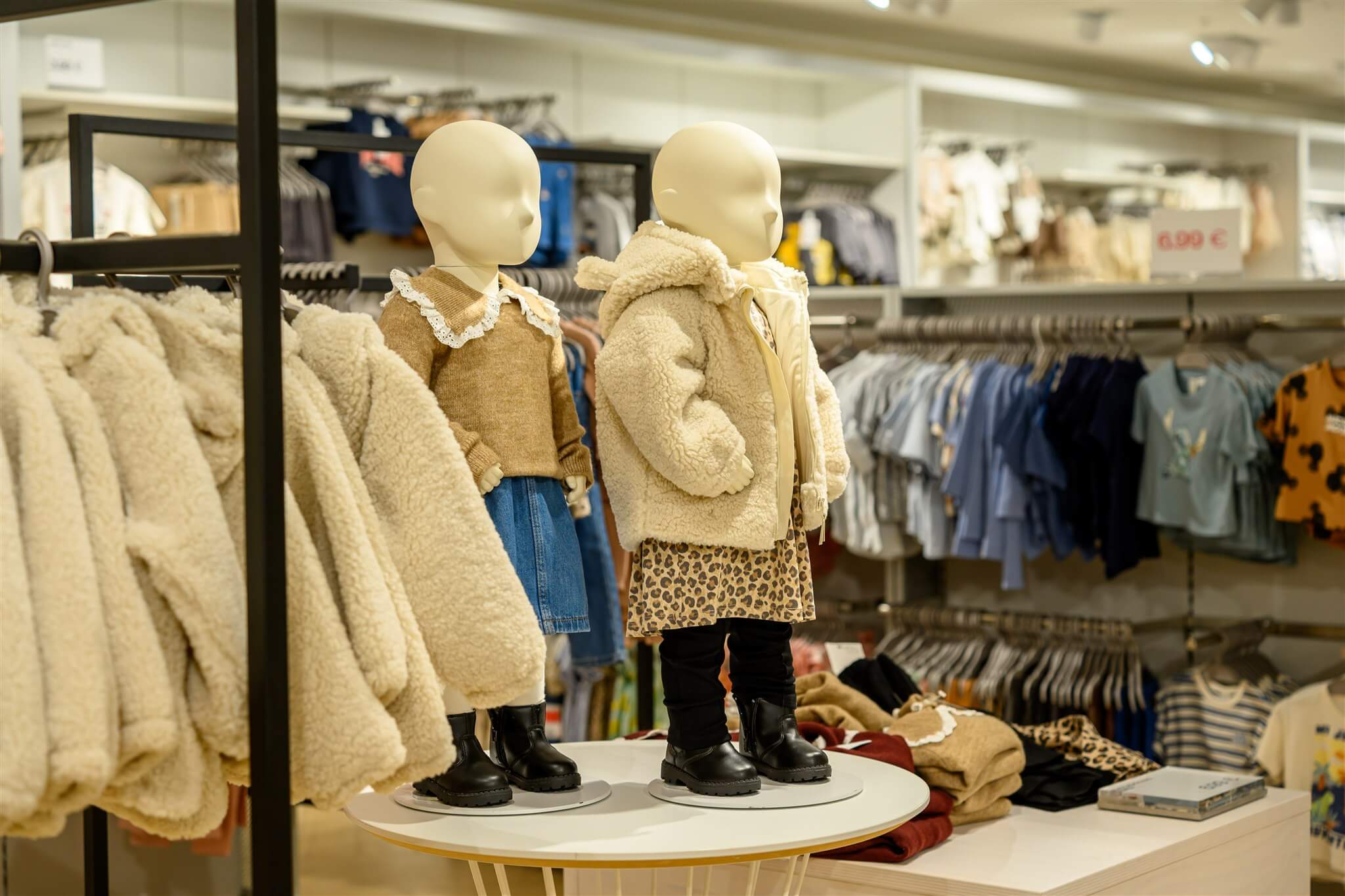 Two child mannequins in a clothing store display winter outfits with fluffy coats and boots, surrounded by racks of children's clothes.