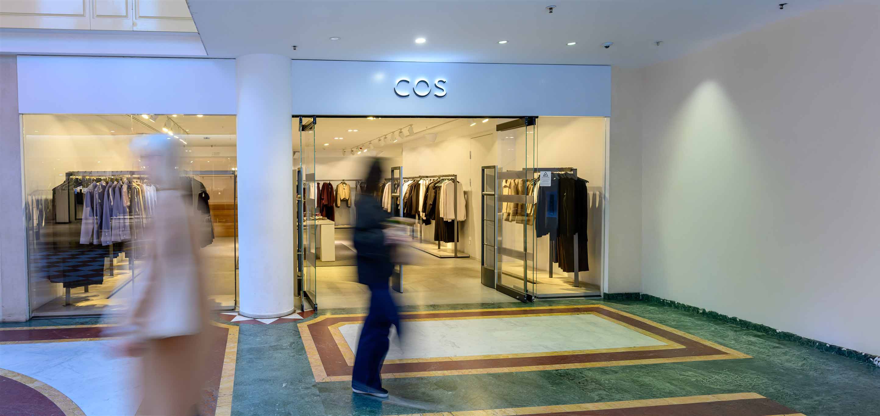 Two blurred figures walk past a COS clothing store with a minimalist interior and neatly arranged garments on racks.