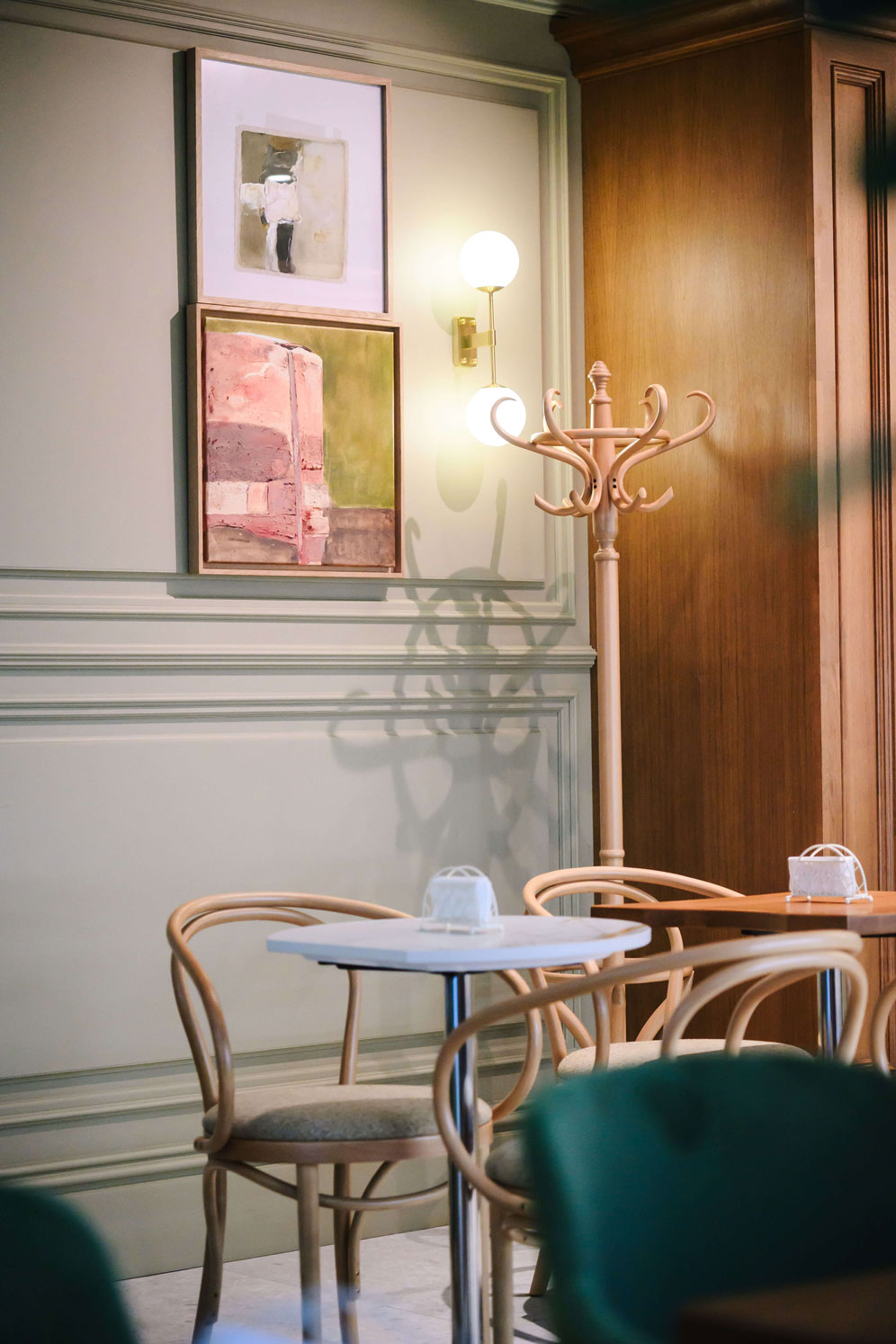 Cozy cafe interior with wooden chairs, small round tables, wall art, and a coat rack. Soft lighting creates a warm atmosphere.