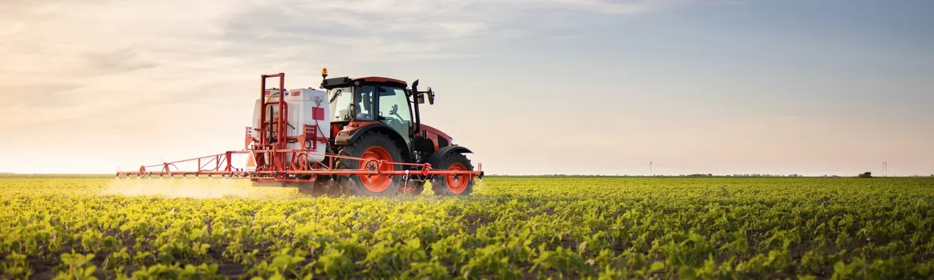 A red tractor sprays crops in a vast green field under a clear sky at sunset.