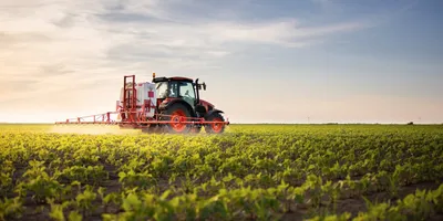 A red tractor sprays crops in a vast green field under a clear sky at sunset.