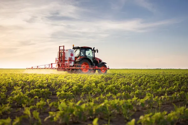 A red tractor sprays crops in a vast green field under a clear sky at sunset.