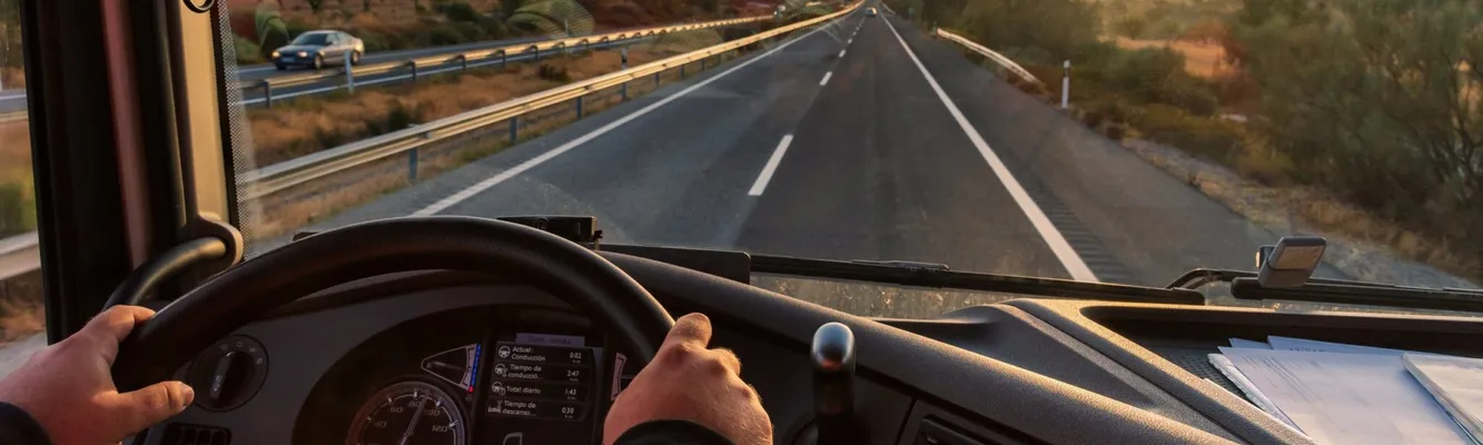 View from a truck cabin showing hands on the steering wheel, driving on an open highway at sunset, with a clear sky and distant hills.