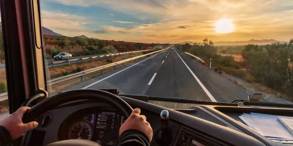 View from a truck cabin showing hands on the steering wheel, driving on an open highway at sunset, with a clear sky and distant hills.