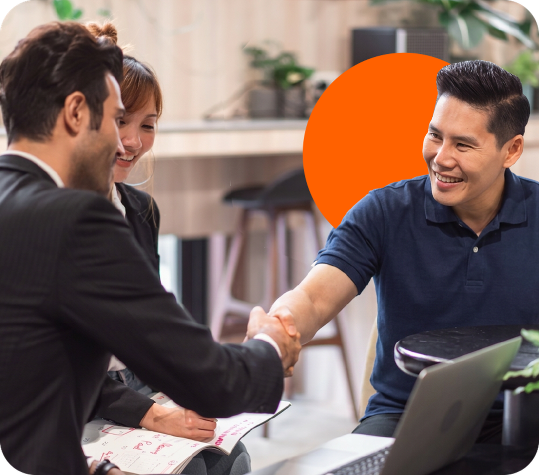 Three professionals shaking hands at a desk with laptops and paperwork, smiling in a modern office setting.