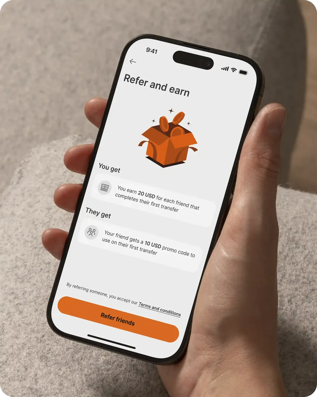 Hand holding smartphone displaying Ria app "Refer and earn" screen with gift box icon and orange "Refer friends" button.
