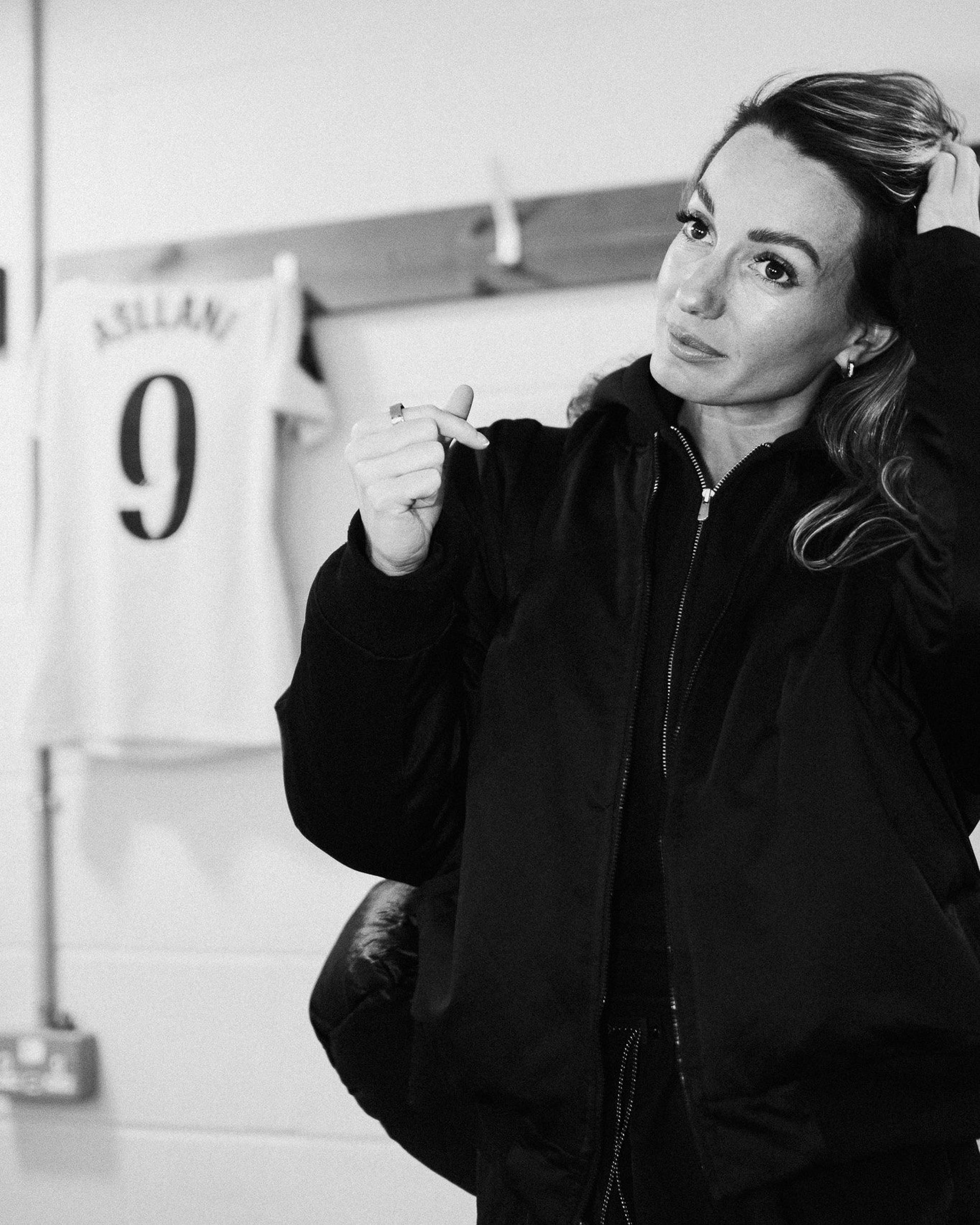 Black and white portrait of a footballer in a changing room, her number 9 shirt hanging behind her