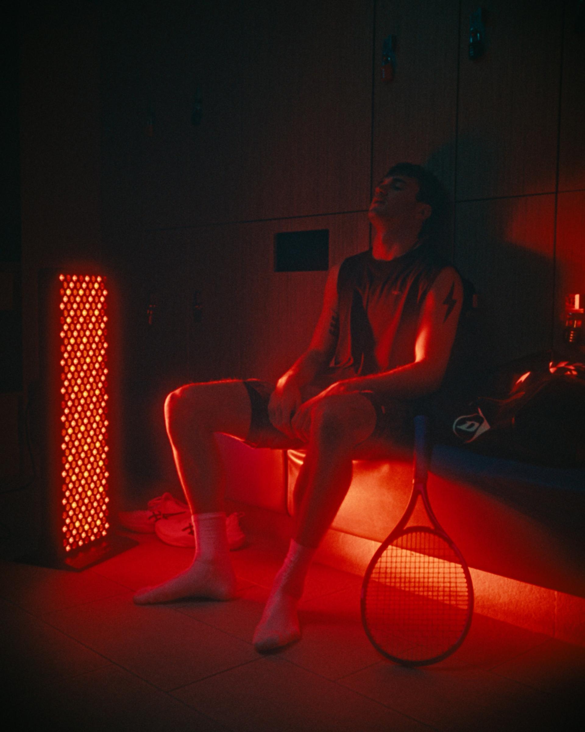 Jack Draper resting on a locker room bench in red light from a Flowlight panel, tennis racket at his side.