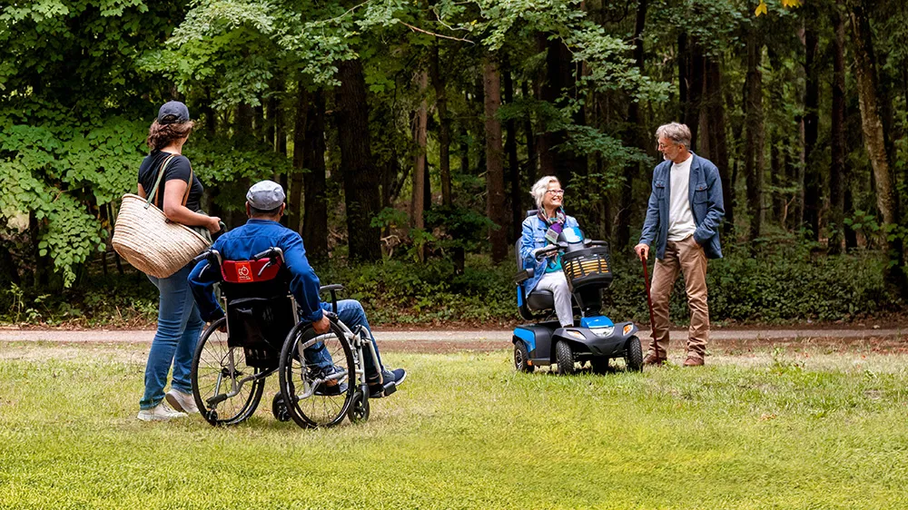 Een groep mensen staat bij elkaar in een park. Ze gebruiken verschillende hulpmiddelen. Een scootmobiel, een rolstoel en een wandelstok.