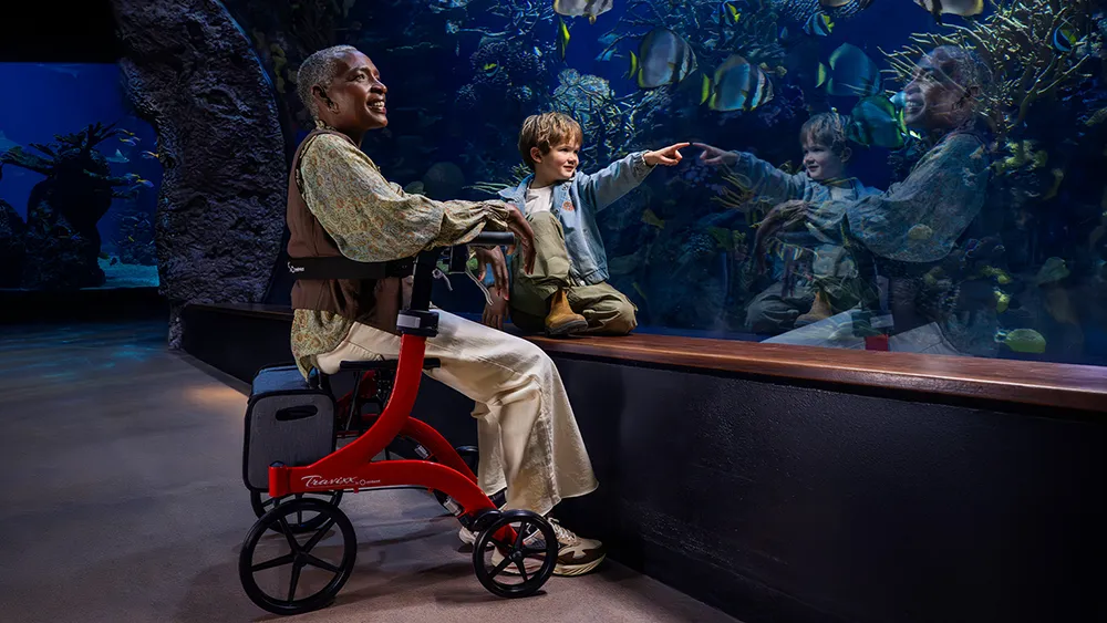 Vrouw en kind samen bij aquarium. Vrouw zit lachend op een rollator.