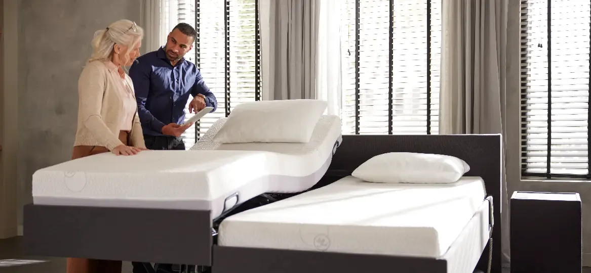 Twee mensen bespreken een verstelbaar bed tijdens advies aan huis, met grote ramen op de achtergrond.