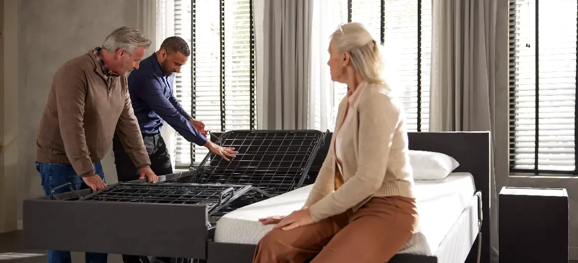 Twee mannen stellen een verstelbaar bedframe af terwijl een vrouw op het matras zit, met zonlicht dat door de nabijgelegen ramen naar binnen schijnt.