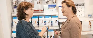 Twee vrouwen staan voor een schap in de apotheek, waar ze informatie over het lenen van medische hulpmiddelen bekijken.