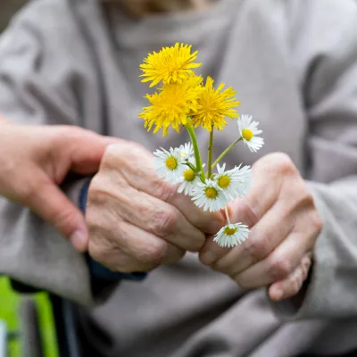 Oudere persoon die witte en gele bloemen vast houdt en een hand van iemand anders op de pols heeft
