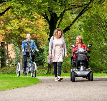 Een vrouw wandelt naast een man en een andere vrouw die op een fiets rijdt, in een levendige buitenomgeving.