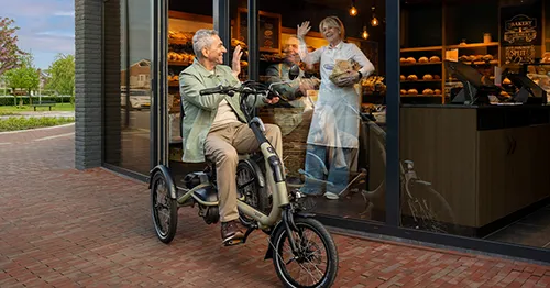 Beeld van een meneer op een driewielfiets. Hij zwaait bij een bakker via het raam naar binnen. De bakker staat binnen en zwaait terug