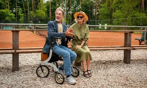 Man en vrouw zitten met rollator bij de tennisbaan