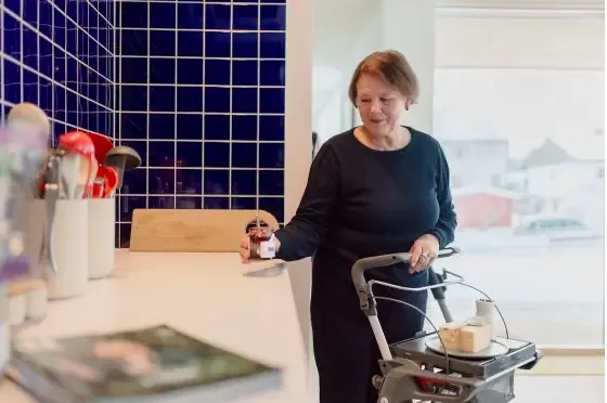 Oudere vrouw gebruikt een binnenrolator van het merk TOPROi in een keuken, ze houdt een potje jam vast. In een blauw getegelde keuken