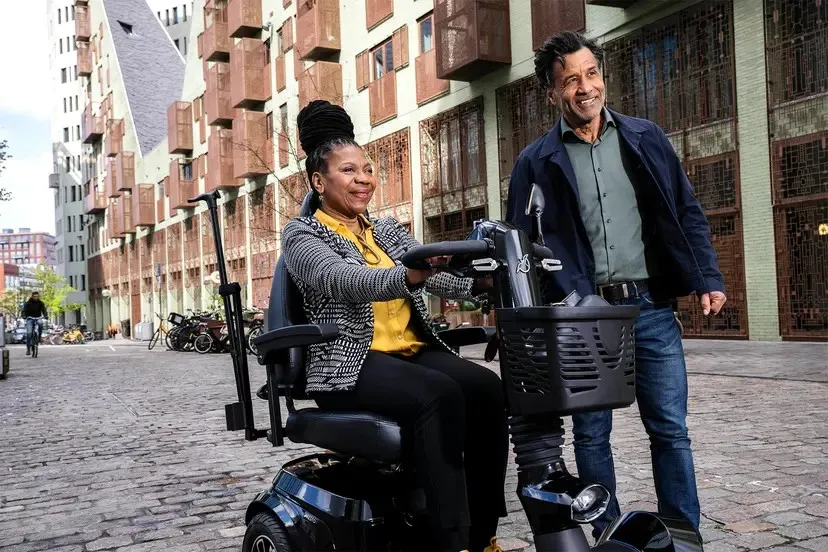Een vrouw rijdt op een scootmobiel over een straat met kasseien, vergezeld door een man die naast haar loopt. Aan weerszijden van de straat staan ​​gebouwen.