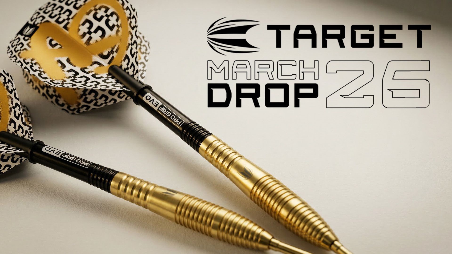 Gold Target darts (Target Bolide Envy) with patterned flight wings, alongside the text "TARGET MARCH DROP 26" on a beige background.