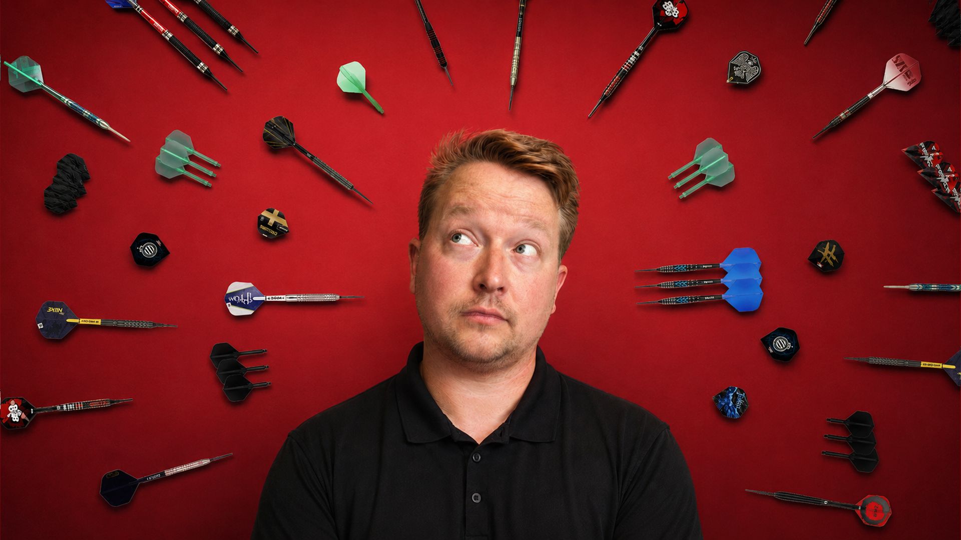 A man in a black shirt looks up thoughtfully at various colorful darts scattered on a red background.