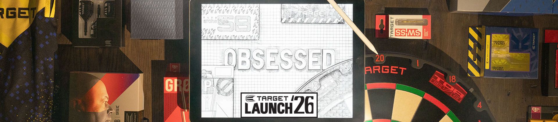 A tablet displaying "Obsessed" and "Target Launch 26" is surrounded by darts-related merchandise and a dartboard on a wooden surface.
