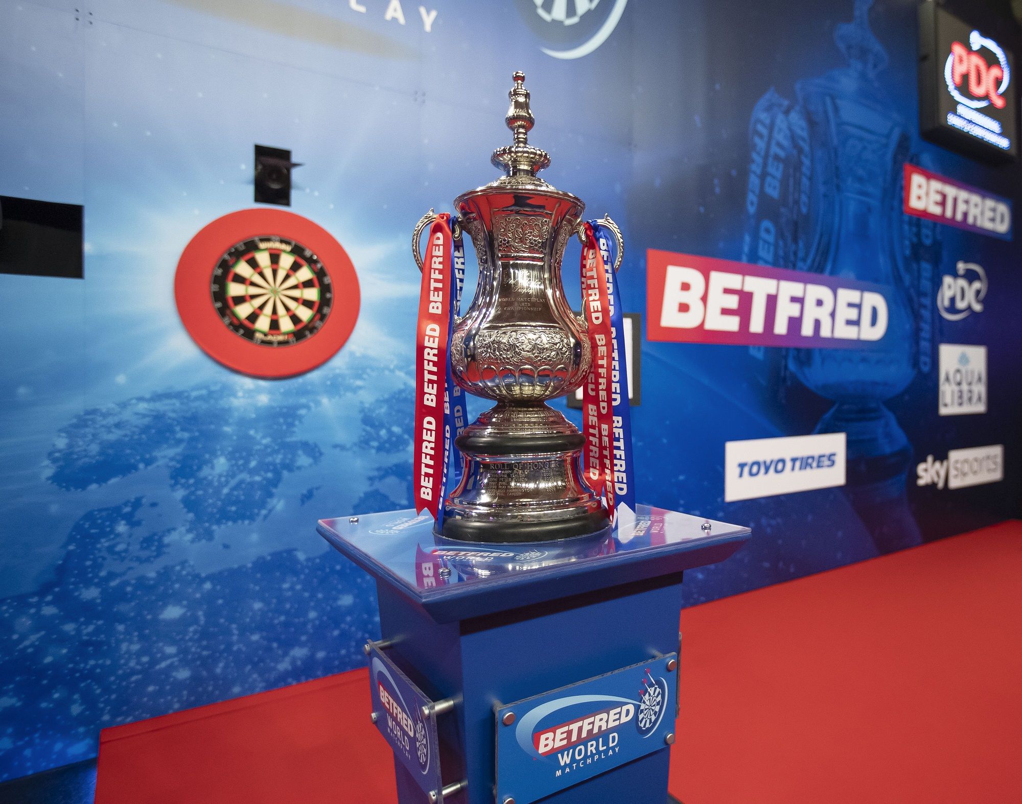 A shiny silver trophy with "Betfred" ribbons stands on a podium in front of a dartboard on a stage with various sponsor logos.