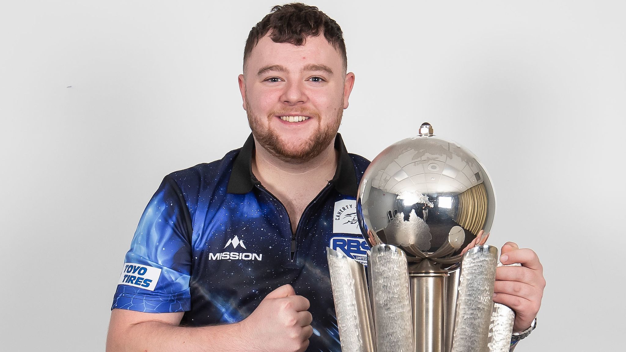 Smiling man in a blue patterned shirt holding a large silver trophy with sphere on top against a white background.