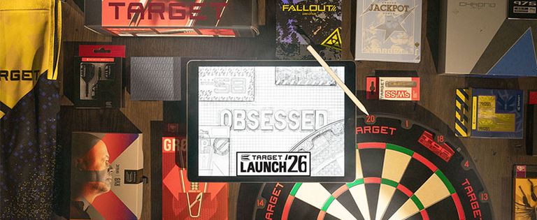 A tablet displaying "Obsessed" and "Target Launch 26" is surrounded by darts-related merchandise and a dartboard on a wooden surface.