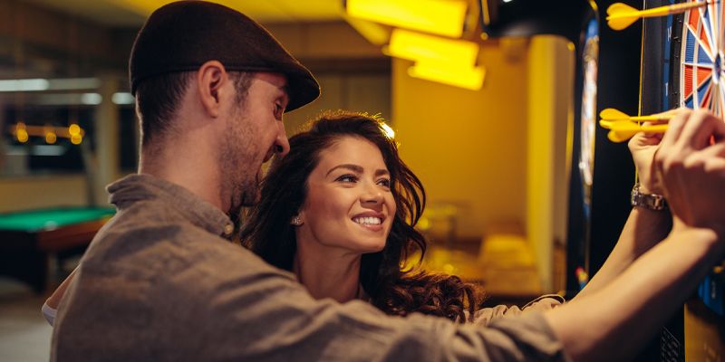 A smiling couple stands in front of a dartboard in a warmly lit game room, with pool table visible in the background.