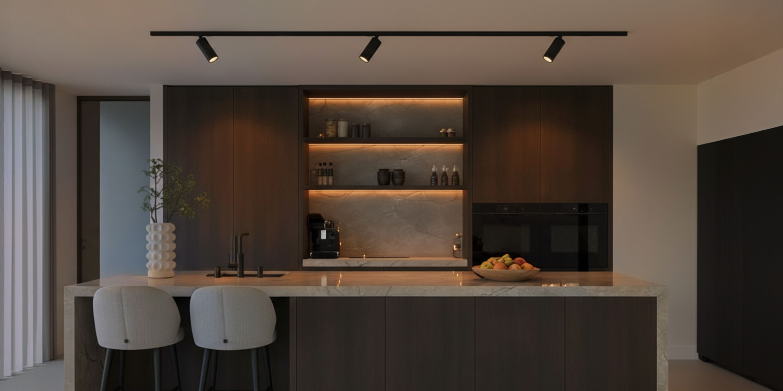 Modern kitchen with dark wood cabinets, marble countertops, and recessed lighting. Features a kitchen island with two chairs and a bowl of fruit.